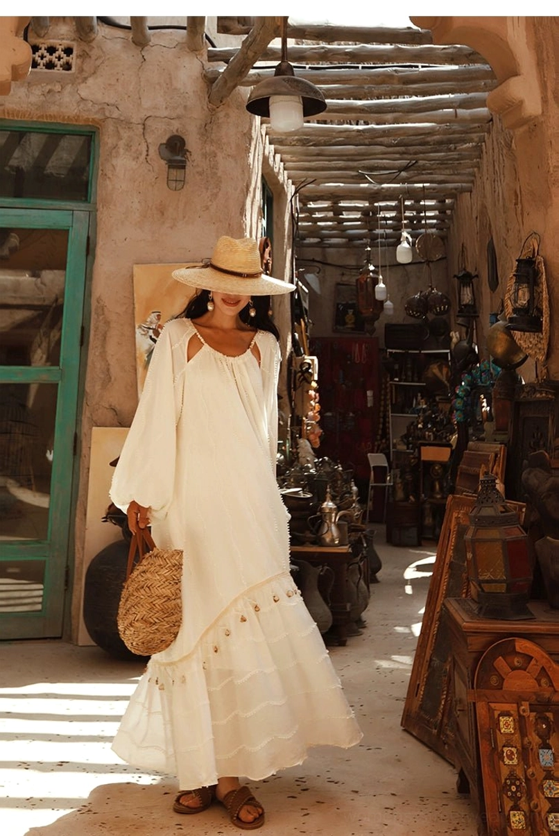 Front view of a white bohemian maxi dress with square neckline, relaxed fit, long lantern sleeves, ruffled hem, straw tote bag, and vacation streetwear styling