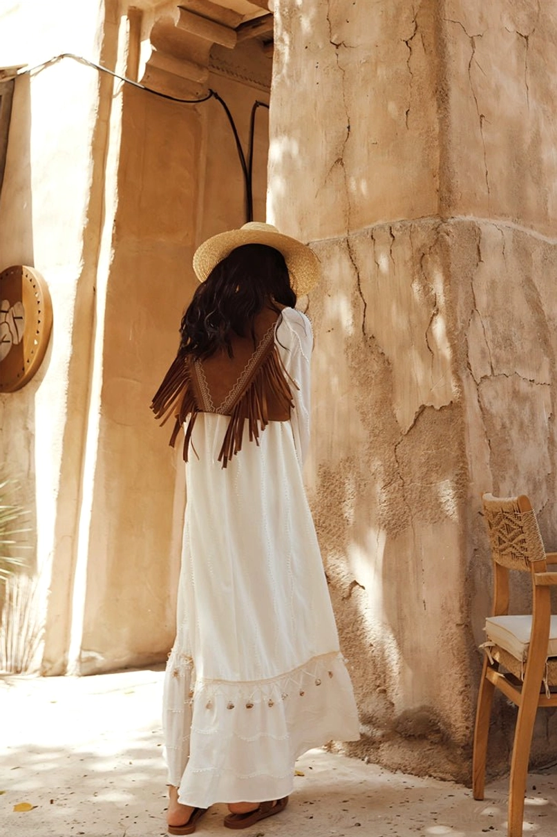 Rear full-body view of a flowing white boho maxi dress layered with a brown fringe vest and styled for desert streetwear fashion