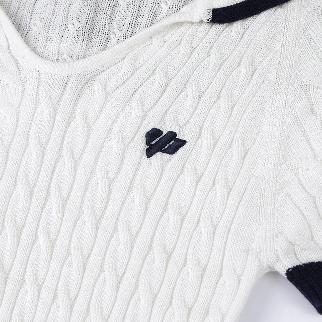 Close-up detail of the textured cable knit fabric, embroidered chest badge, and navy blue contrast edging on a cream white cropped sailor collar top