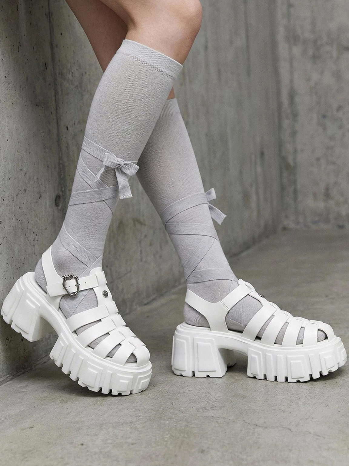 Side view of white chunky fisherman platform sandals worn with grey bow-detail socks against an industrial concrete wall