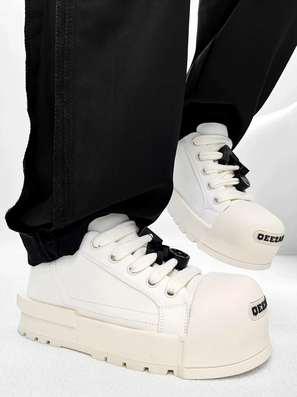 White chunky platform sneakers with black laces and decorative lace charms worn with black straight leg trousers