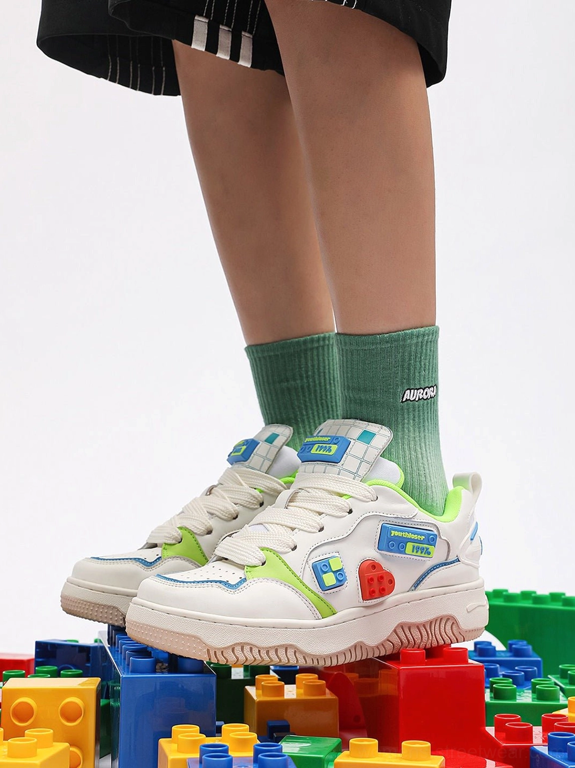 White chunky platform skate sneakers styled with green socks and black shorts showing the padded upper colorful patches and thick treaded outsole