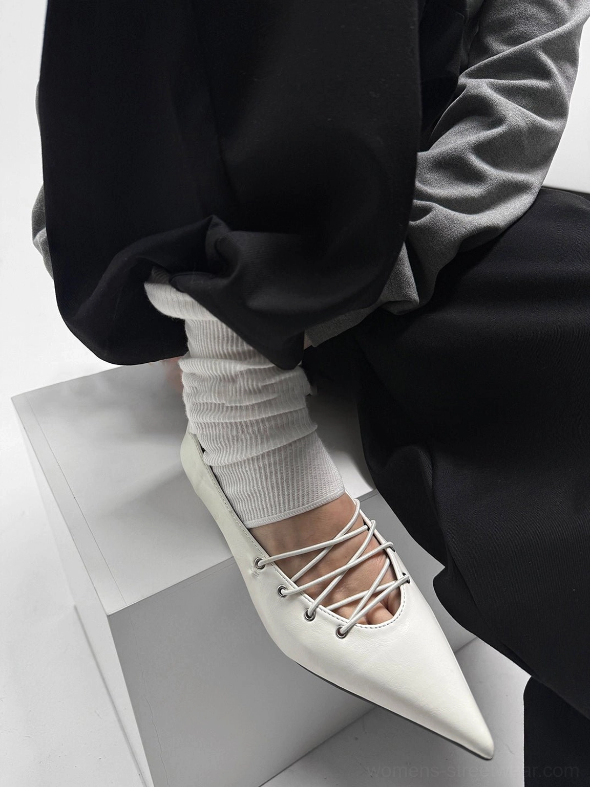 Model wearing white corset-lace pointed ballet flats with white socks, black relaxed trousers, and a grey top in a minimalist streetwear outfit