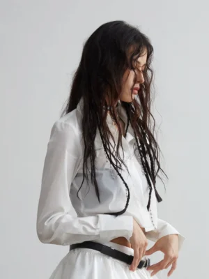 Side back view of a white cropped cotton long-sleeve shirt styled with a ruffled mini skirt and black leather belt