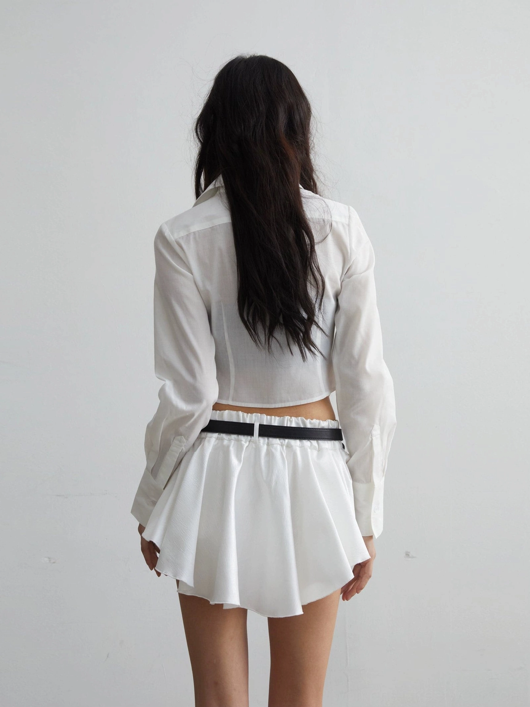 Back view of a white cropped long-sleeve shirt paired with a low-rise ruffled mini skirt and black waist belt