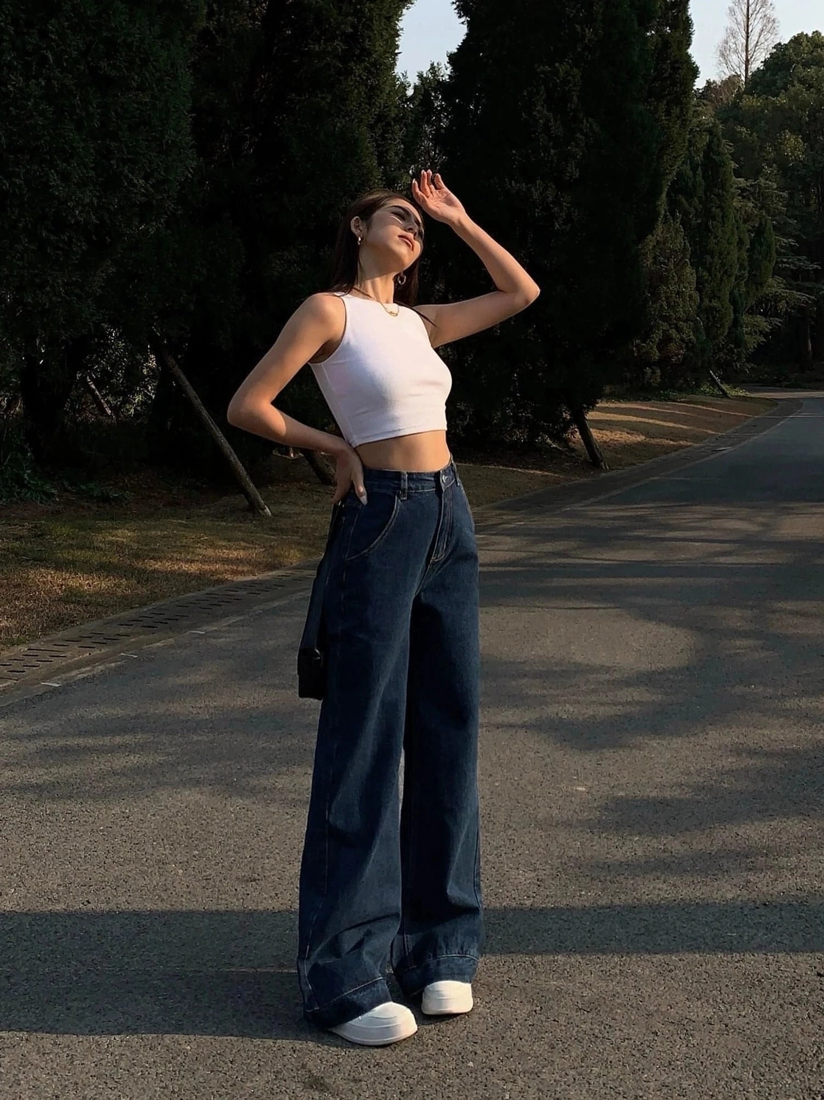 White cropped tank top back side full body shot - minimalist sleeveless cropped design showing waistline paired with dark blue wide-leg jeans and white sneakers on tree-lined outdoor road