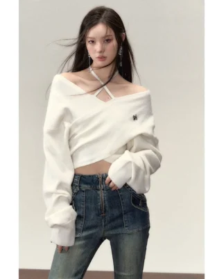 Model wearing a white cross wrap knit top with crisscross halter straps, off shoulder neckline, cropped hem, chest brooch and relaxed bishop sleeves with high waisted denim