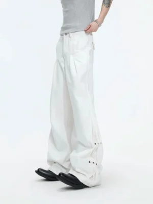 White draped wide-leg streetwear pants shown from a low side angle with pleated waist, flowing cotton silhouette, lower-leg metal rivets, and adjustable snap details