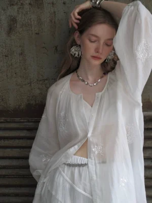 Front view of a white embroidered peasant blouse set with a button-front neckline, floral beadwork, lightweight fabric, and romantic lantern sleeves styled against a retro industrial wall