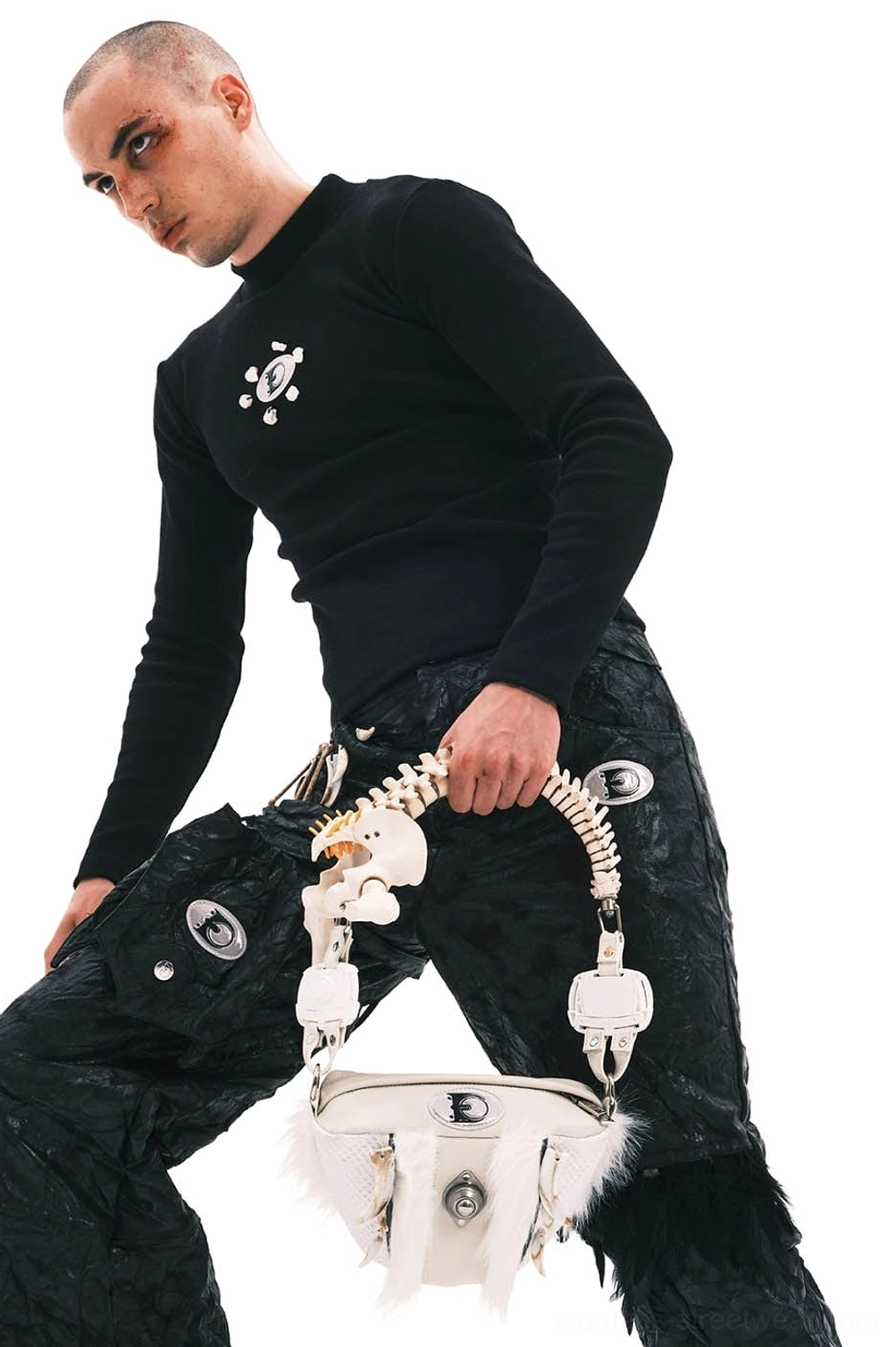 Male model holding white faux fur skeletal spine handle shoulder bag with black cargo pants in avant-garde streetwear look
