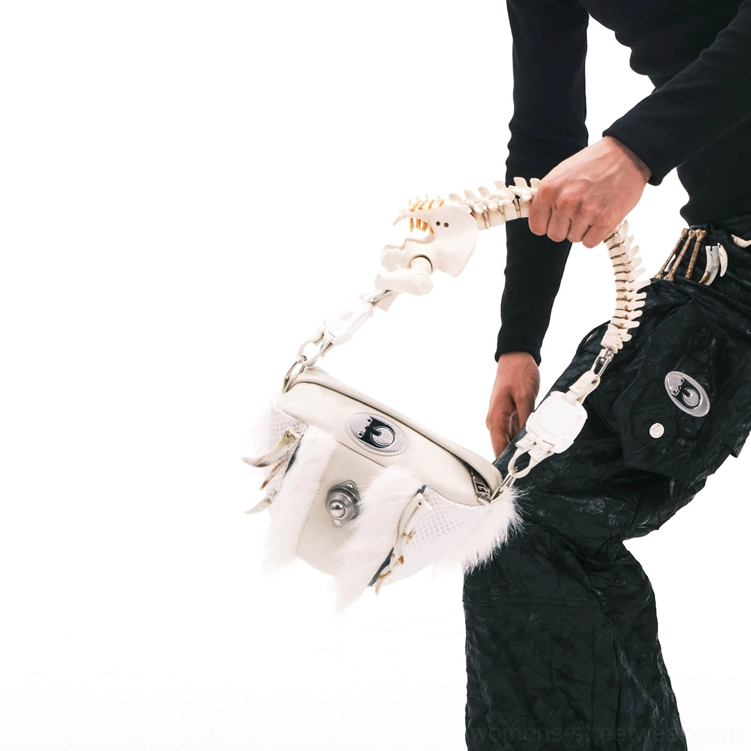 White faux fur skeletal spine handle shoulder bag held above black tactical cargo pants, showing cream leather body and silver hardware