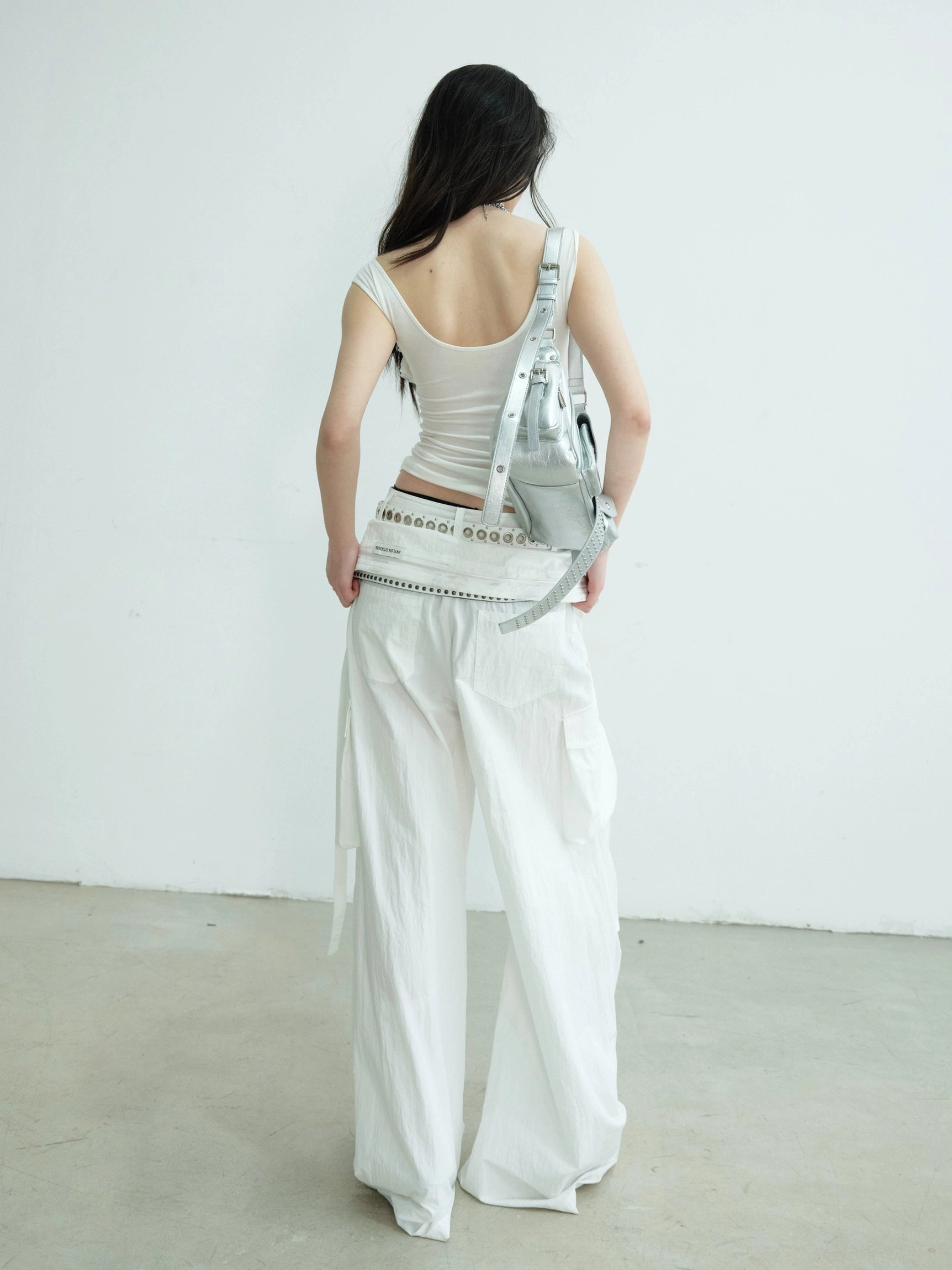 Back view of white fitted sleeveless tank top styled with studded belt white cargo pants and silver backpack