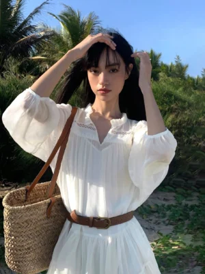 Close-up of white French-inspired mini shirt dress with lace-trimmed V-neck, flowing pleated skirt, brown belt and straw shoulder bag