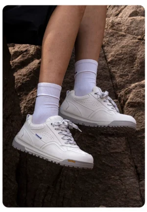 White casual outdoor sneakers worn with crew socks on rocky terrain, featuring perforated side panels, subtle gray accents, and chunky rubber soles