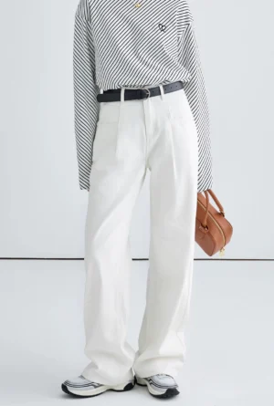 Women wearing white high-rise wide-leg streetwear jeans with a striped long-sleeve top, black leather belt, tan handbag, and two-tone sneakers in a minimalist studio