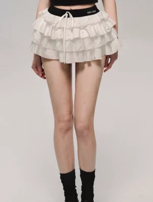 Close-up detail of white lace tiered mini skirt lower body showing multi-layer ruffle hem, branded elastic waistband, and front drawstring design