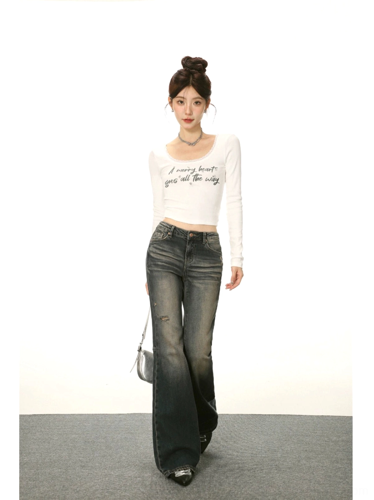 Full-body styling view of a woman wearing a white lace trim cropped long-sleeve top with vintage distressed flared jeans and a gray underarm bag