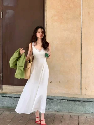 White lace trim camisole maxi dress with fitted waist and flowing ankle length skirt styled with red strappy sandals on a vintage street