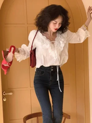 White lace-trimmed ruffle blouse with V-neck tie detail and flared cuffs styled with high-waisted dark flare jeans in a retro indoor setting
