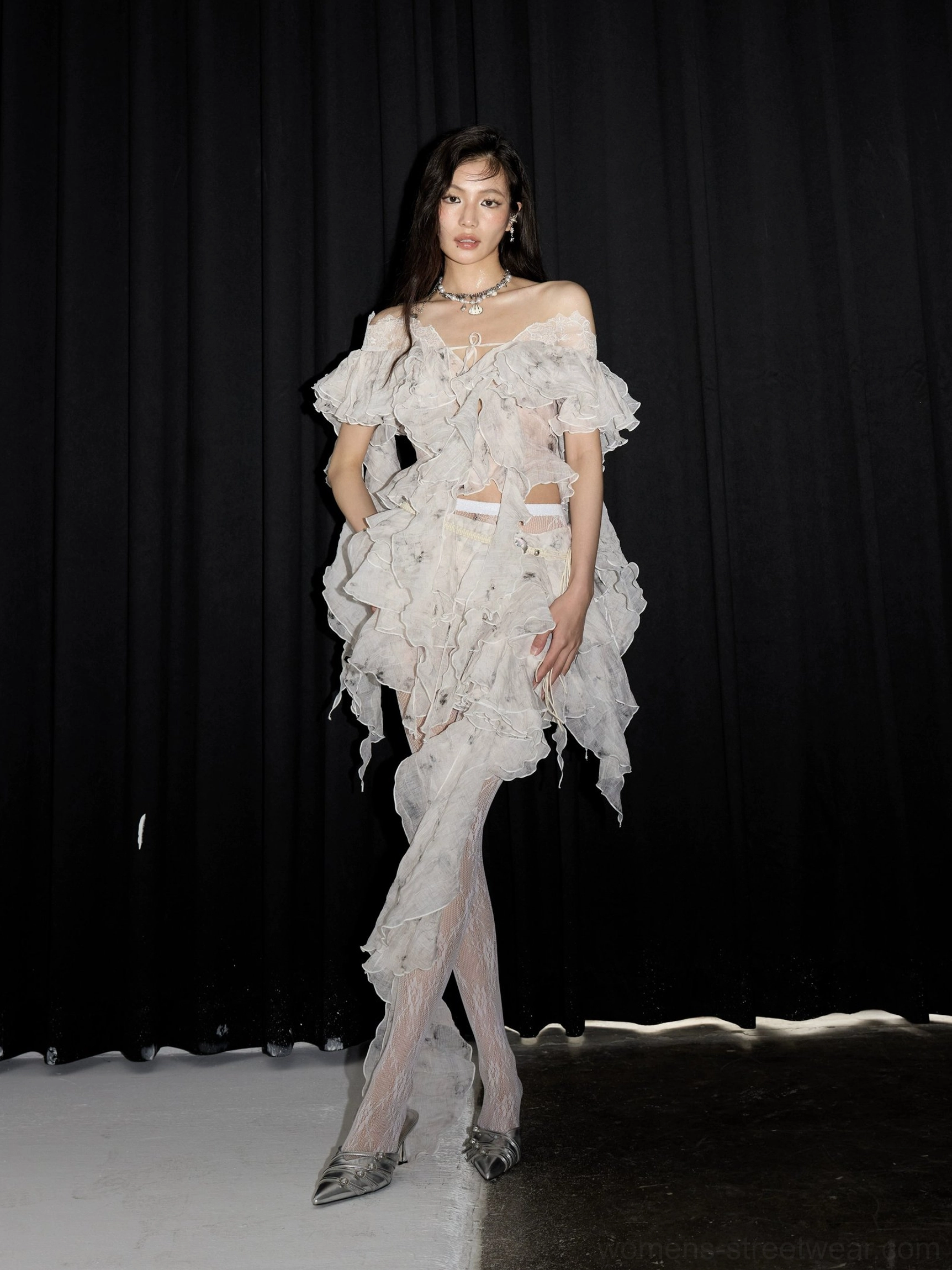Side view of a white off-shoulder lace ruffle two-piece set featuring an asymmetrical draped skirt hem and sheer patterned tights