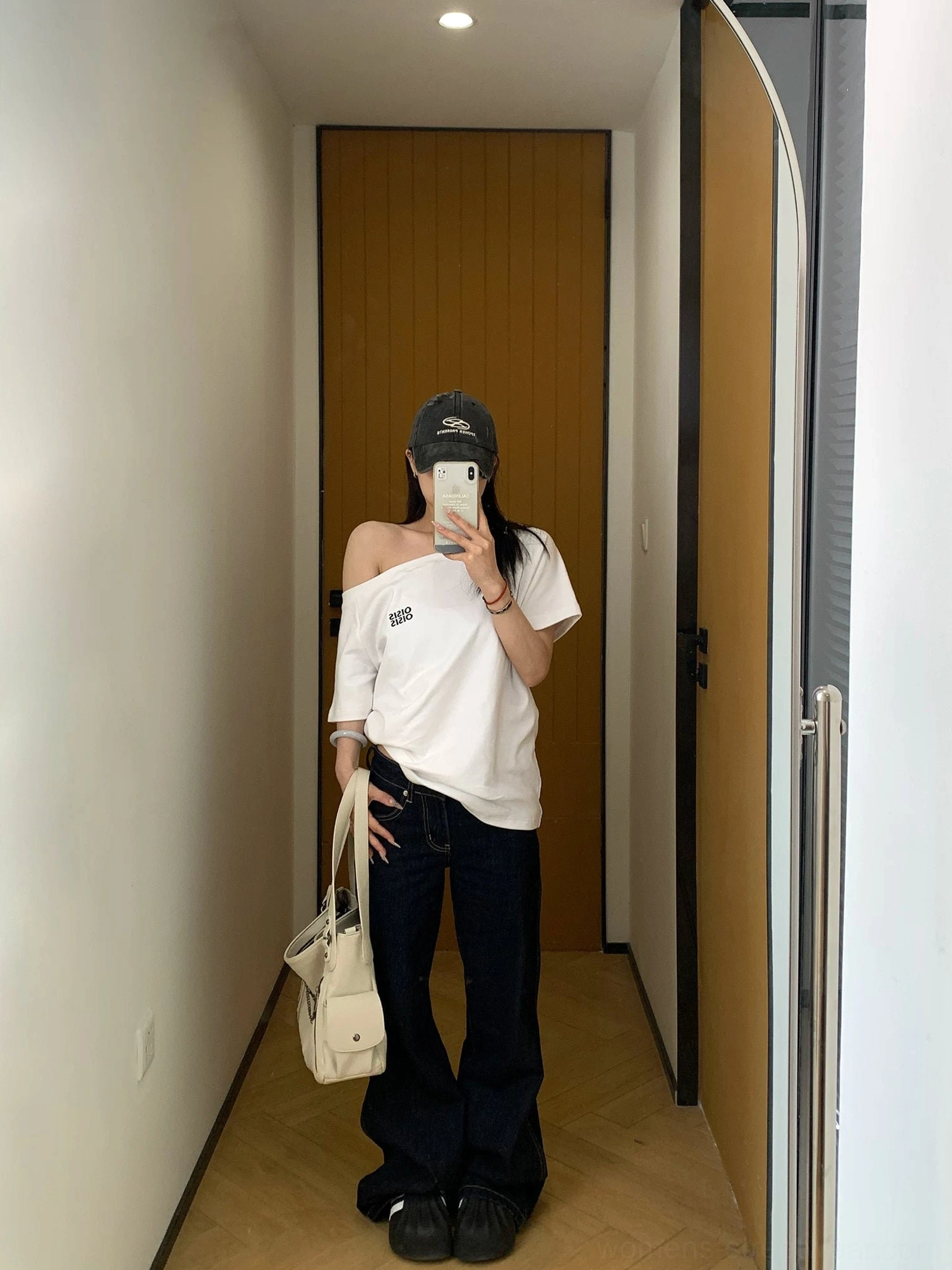 Full-length back view of a minimalist white asymmetrical off-shoulder tee paired with dark flared jeans and a beige handbag