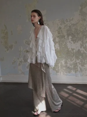 White oversized ruffled chiffon blouse with V-neck, layered front details, lantern sleeves, and metallic wide-leg pants in a vintage sunlit interior