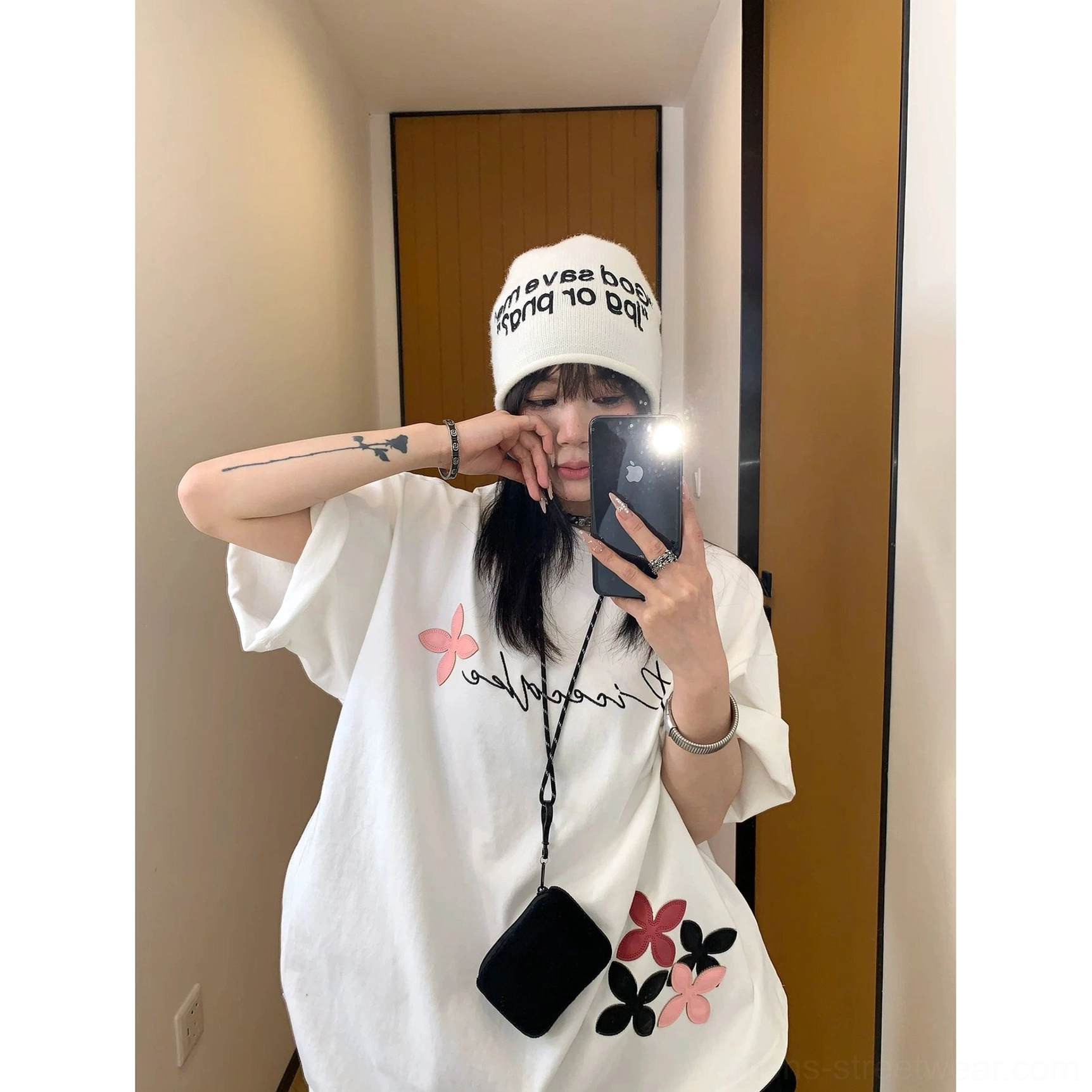 Front styling view of a white oversized script graphic T-shirt with rolled sleeves, floral hem artwork, white beanie, and mini crossbody bag