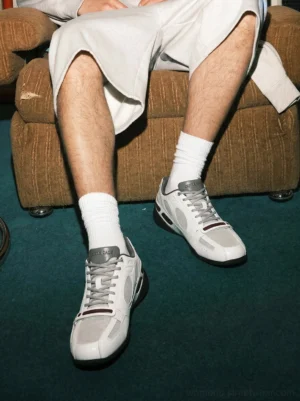 White perforated retro sport sneakers worn with white crew socks and light grey shorts in a casual indoor streetwear outfit