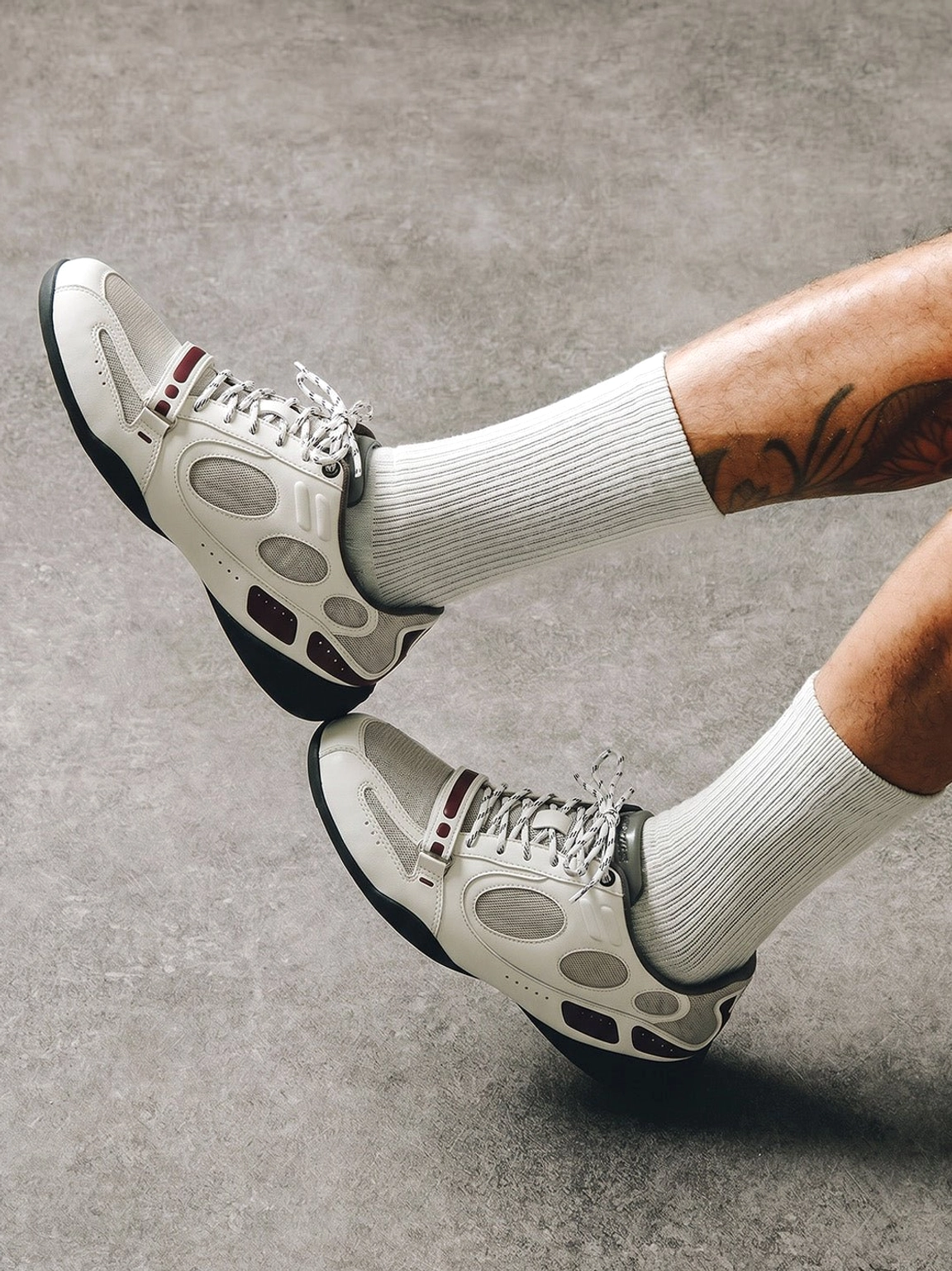 On-foot view of white perforated retro sport sneakers with burgundy details styled with crew socks on a concrete floor