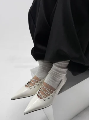 White pointed ballet flats with exposed crisscross laces and glossy upper styled with white ribbed socks and loose black streetwear trousers