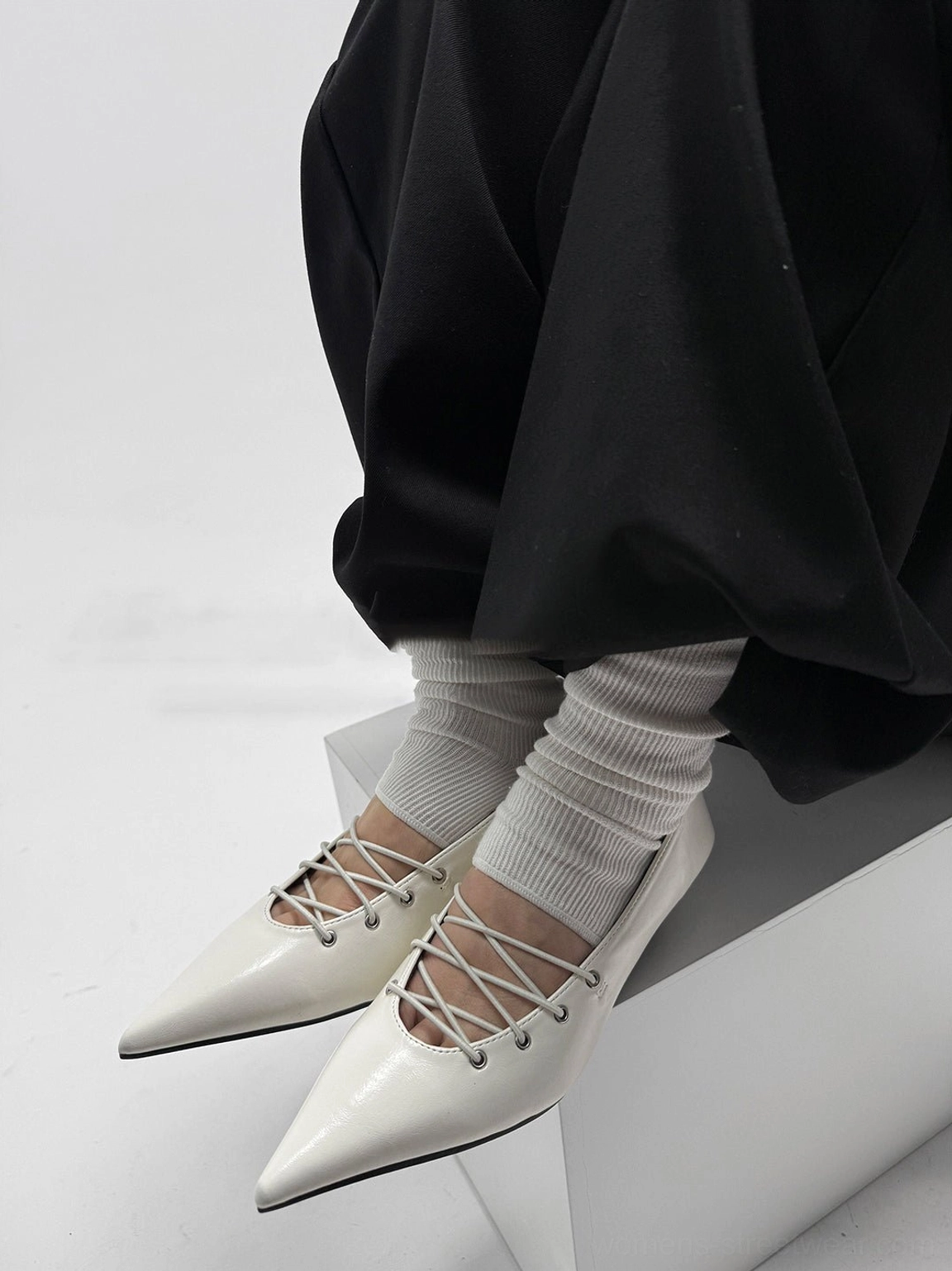 White pointed ballet flats with exposed crisscross laces and glossy upper styled with white ribbed socks and loose black streetwear trousers