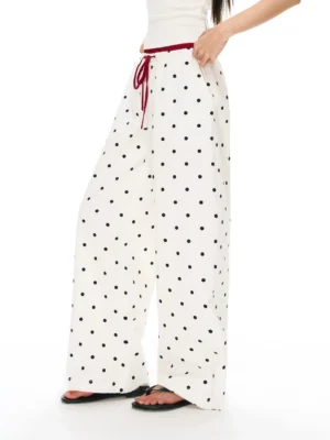White polka dot drawstring wide leg streetwear pants shown from a low side angle with contrasting red waist cord and black slide sandals