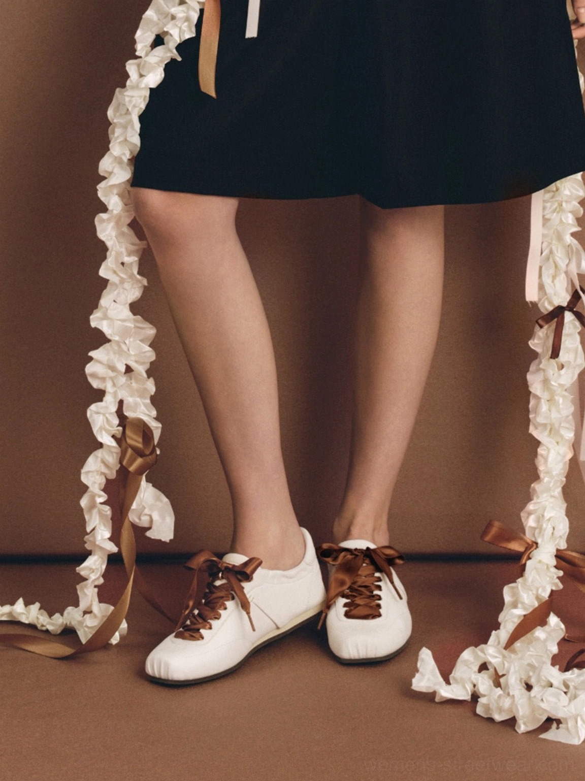 White low-profile retro trainers with brown satin ribbon laces styled with a black mini skirt and feminine streetwear outfit