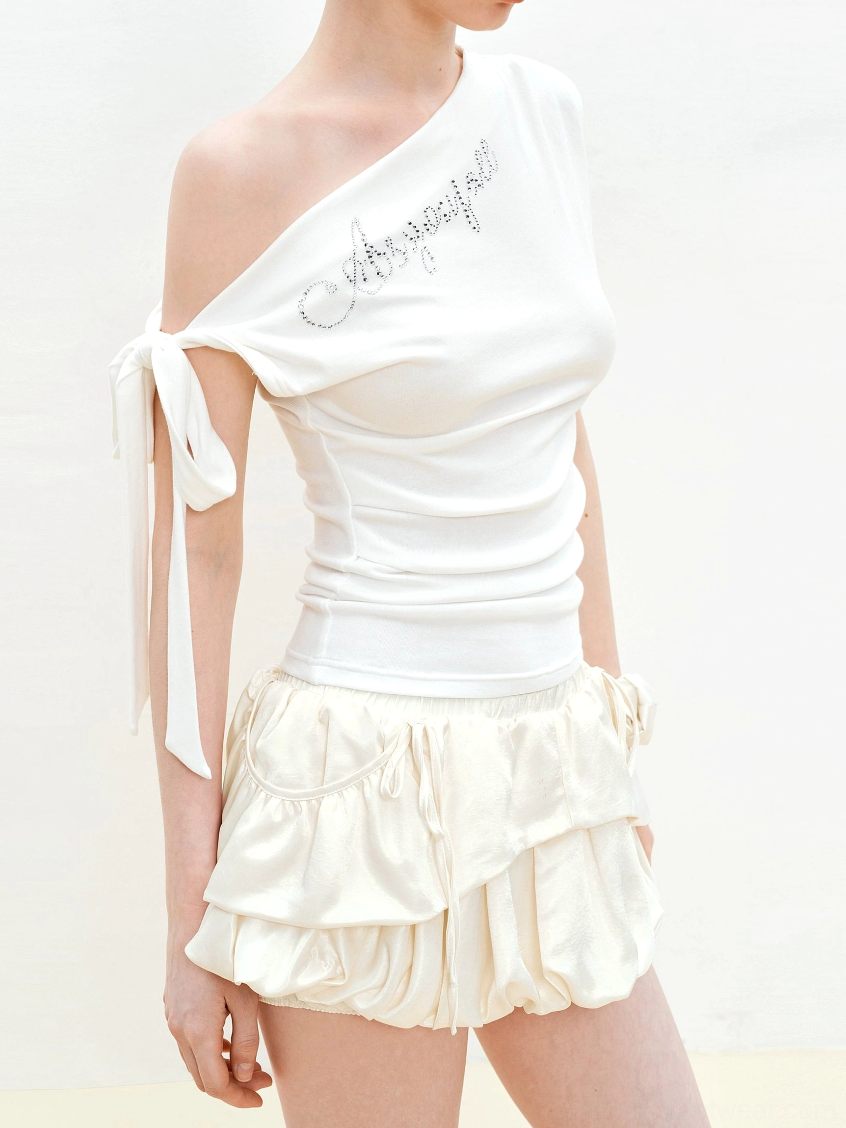White rhinestone one-shoulder bow top with ruched waist shown in a side view with cream ruffle shorts