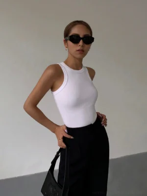 White ribbed knit tank top worn with black suit pants and black handbag, front half-body view on female model wearing black sunglasses, minimalist studio background