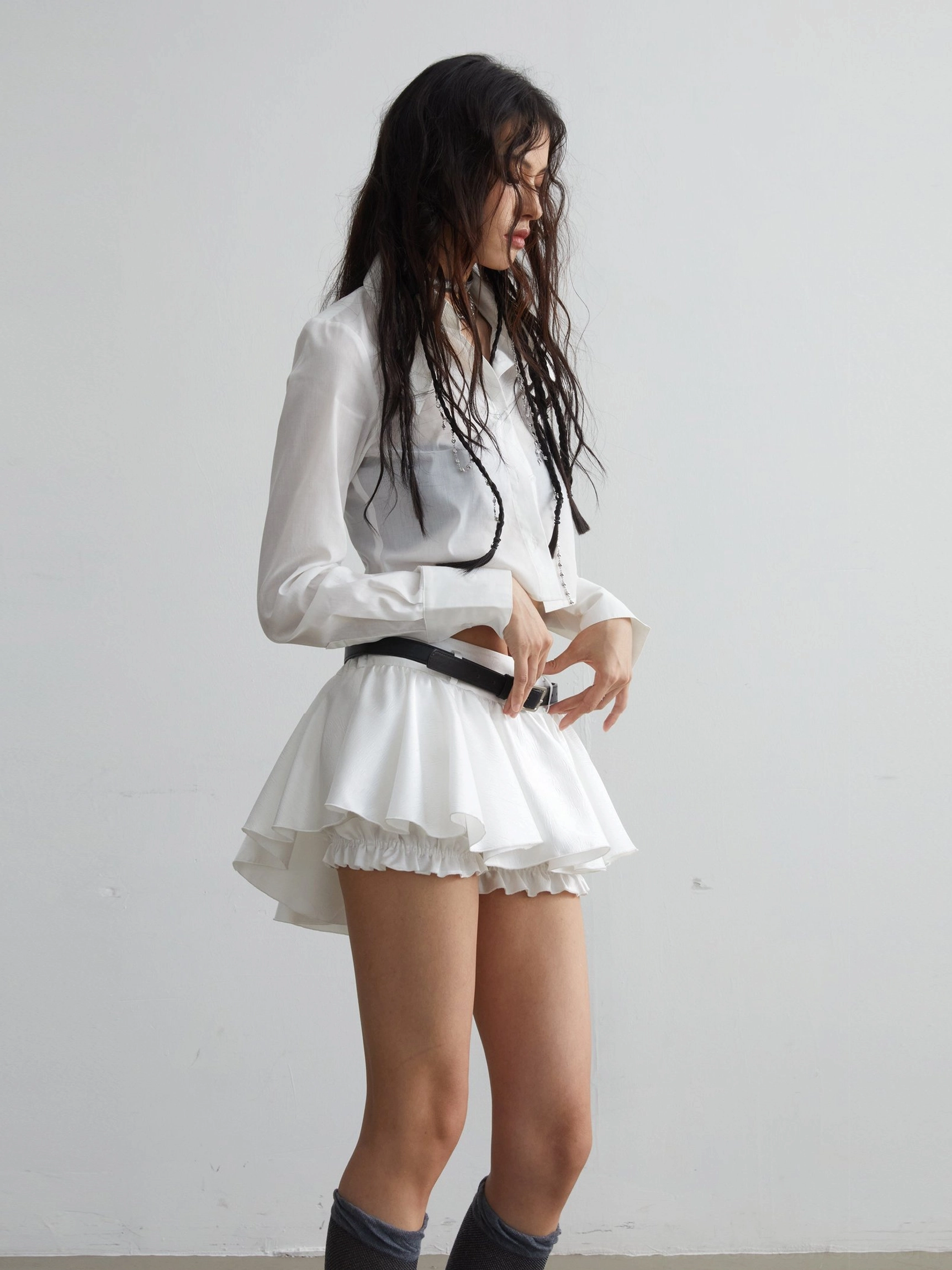 Rear side view of high waisted white ruffled mini skirt with black belt embellishment and cropped shirt