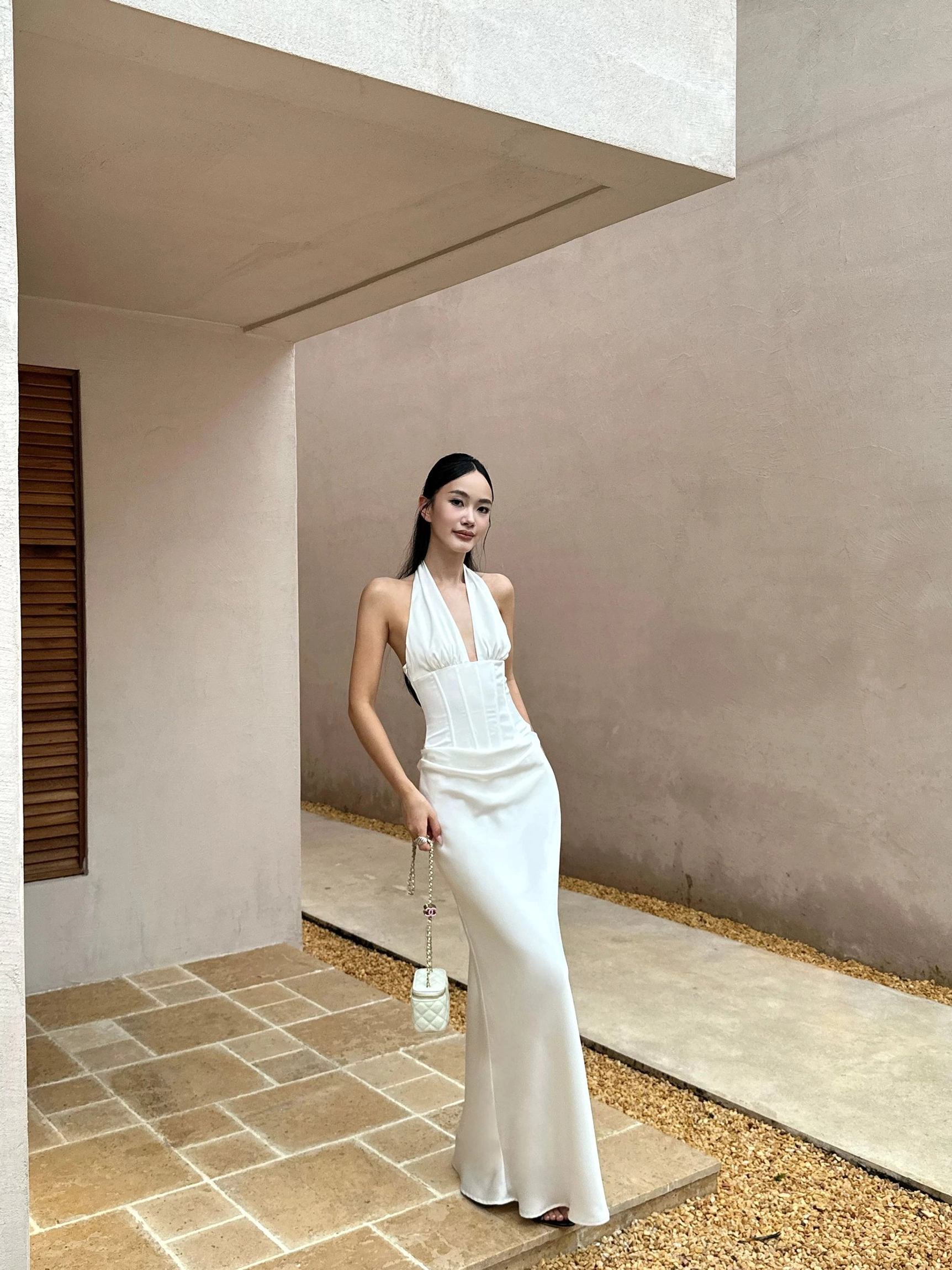 White satin backless maxi dress with halter neckline and ruched waist detail styled with a white chain handbag in an elegant outdoor corridor