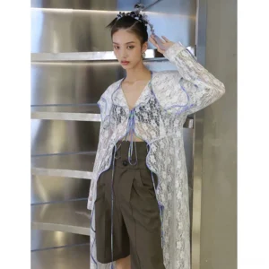 Front full body view of a white sheer floral lace sailor collar duster coat with iridescent blue ruffle trim, adjustable front tie, long sleeves, and flowing asymmetric hem styled with olive tailored shorts