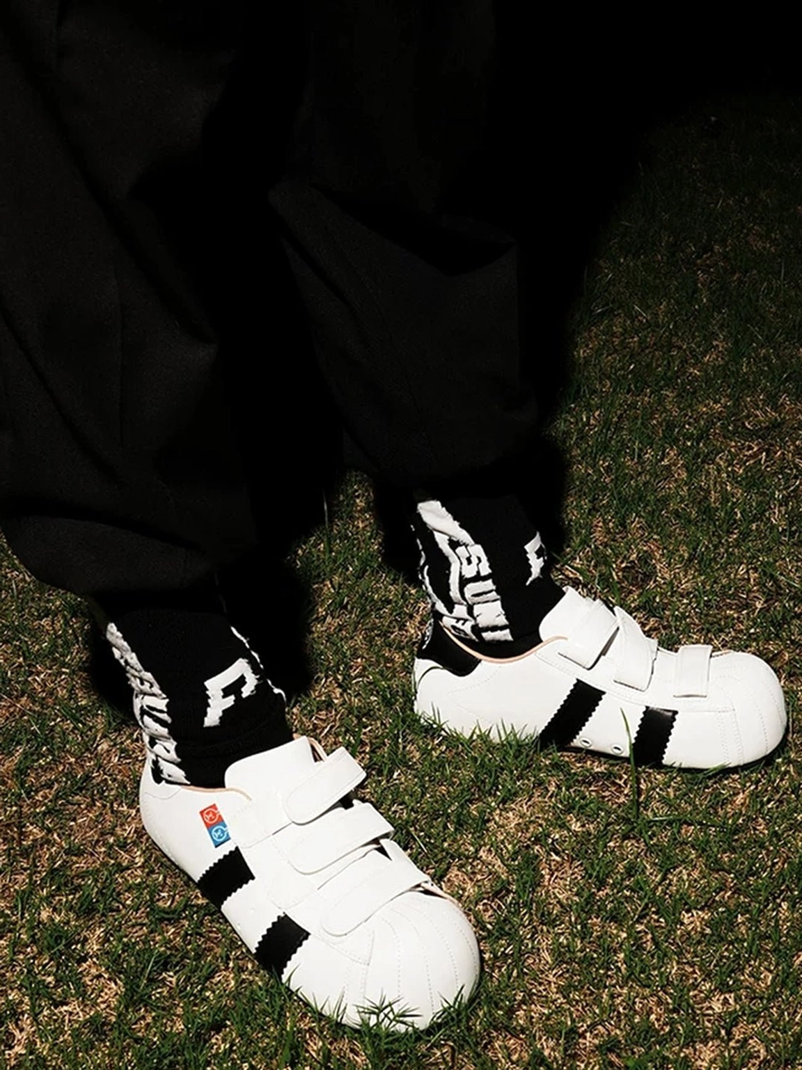 White shell toe hidden lift sneakers with hook-and-loop straps styled with black streetwear pants and graphic socks outdoors