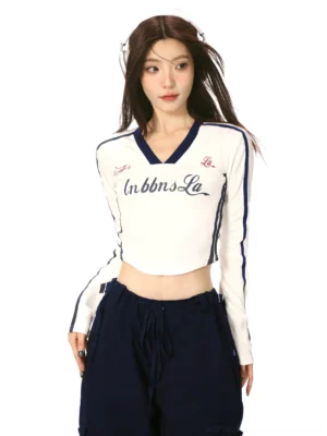 Front view of a white slim fit cropped V-neck long sleeve top with navy contrast neckline, striped sleeves, chest lettering, and Y2K sporty details
