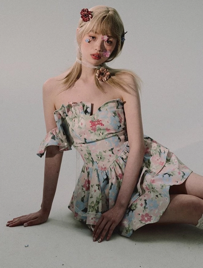 Low-angle seated view of a white strapless floral ruffle mini dress with fitted corset bodice, double-layer mini skirt, and matching watercolor print