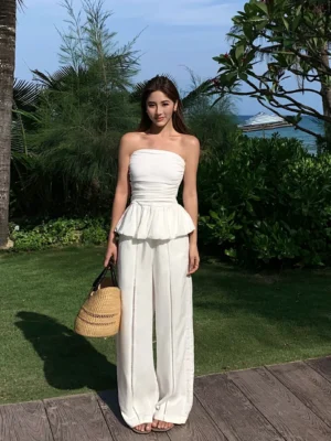 White strapless peplum bandeau top styled with white wide-leg trousers and woven straw tote on a seaside lawn