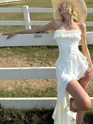Side view of a fitted white strapless maxi dress with layered ruffles and a flowing high slit skirt beside a ranch fence
