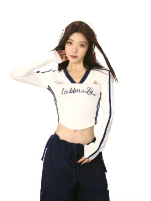 White women's V-neck cropped long sleeve streetwear top with navy color block trim, sleeve stripes, retro chest lettering, and sporty Korean fashion styling