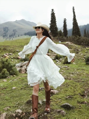 White western ruffle midi dress with billowy long sleeves styled with a cowboy hat sunglasses and brown knee high boots in an alpine grassland setting
