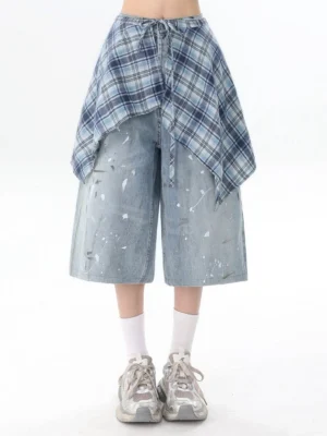 Straight-on view of wide-leg cropped blue denim culottes with asymmetric plaid wrap overlay and tied waist detail styled with white socks and gray chunky sneakers