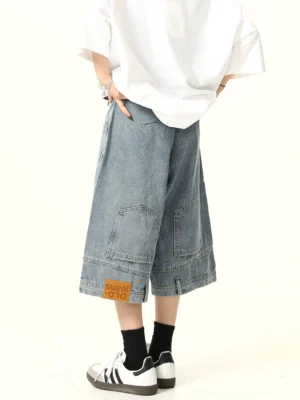 Rear side view of light blue wide-leg denim culotte shorts with oversized rear cargo pockets, hem fabric patch, oversized white T-shirt, black socks, and two-tone sneakers