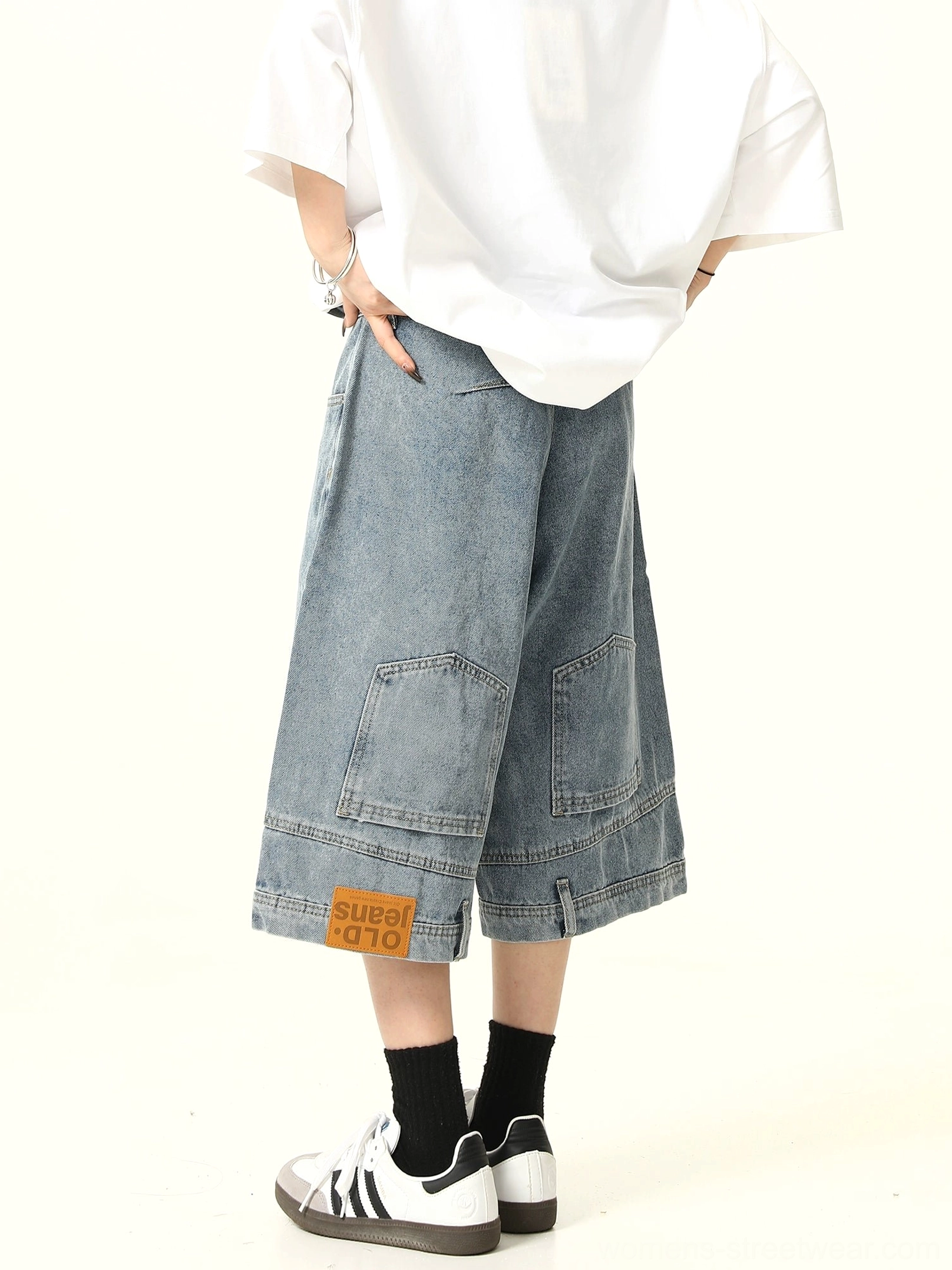 Rear side view of light blue wide-leg denim culotte shorts with oversized rear cargo pockets, hem fabric patch, oversized white T-shirt, black socks, and two-tone sneakers