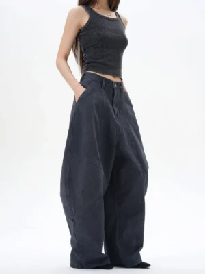 Female model wearing washed black wide leg pleated denim streetwear pants with a high waist and oversized curved seam silhouette in a minimalist white studio