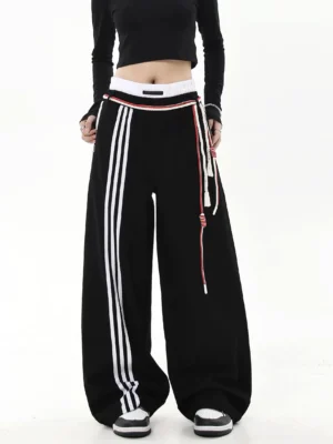 Black oversized varsity track pants front view showing white triple side stripes elastic waistband and rope drawcord details